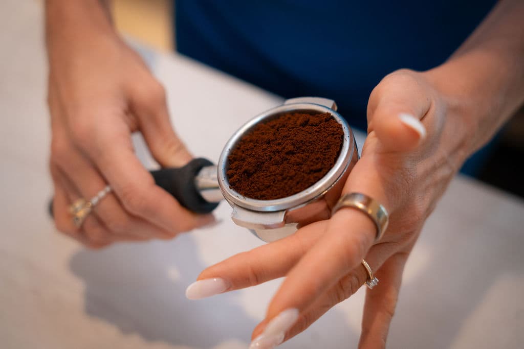 Siebträgermaschine mit eingesetztem Aufsatz zum Zubereiten von Kaffee, vor weißem Hintergrund. Eine Frau mit gepflegten, schönen Fingernägeln bereitet den Kaffee zu.
